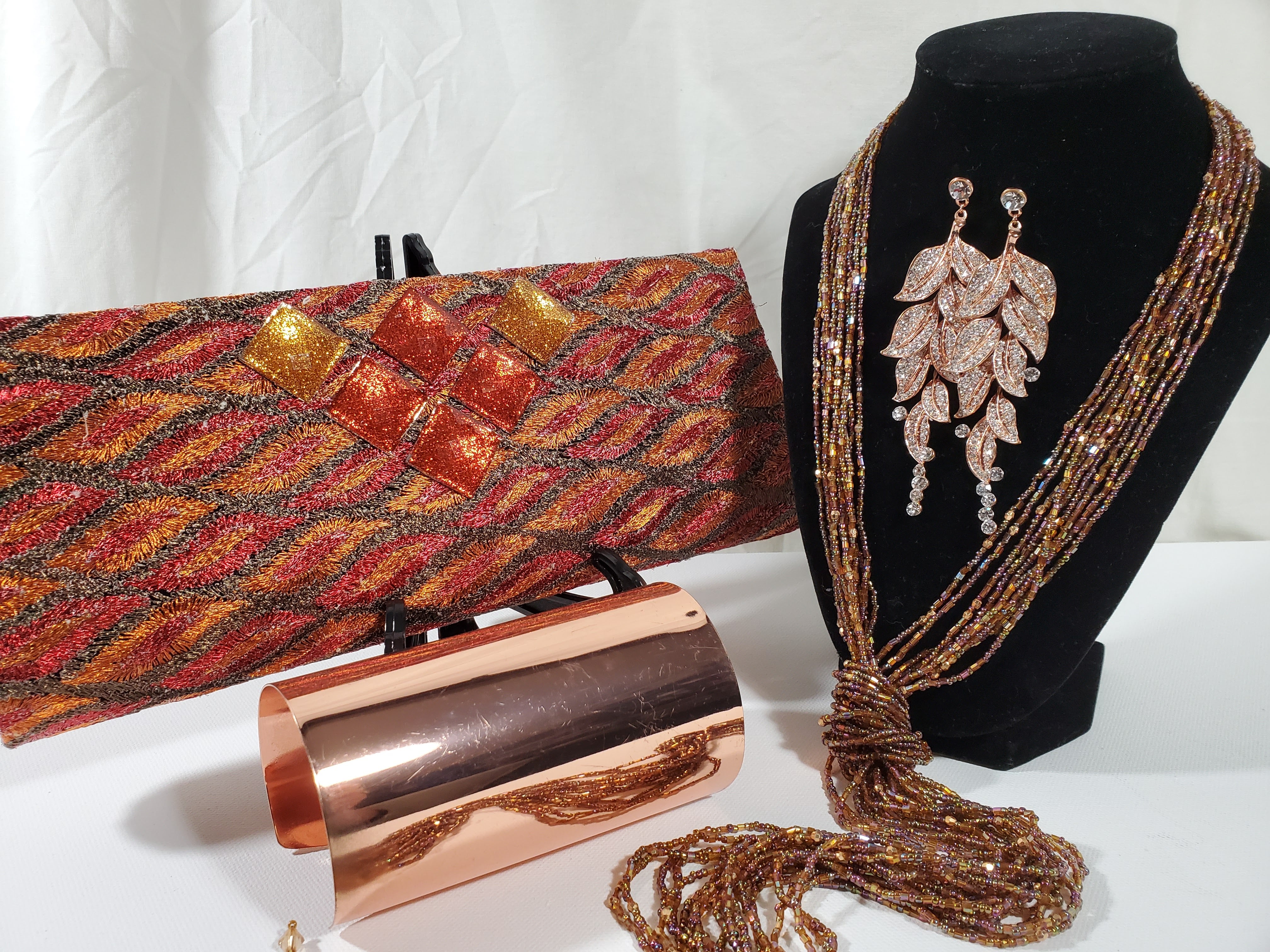 shimmery orange clutch with lasso bead necklace, rose gold rhinestone earrings on stand and copper cuff at forefront